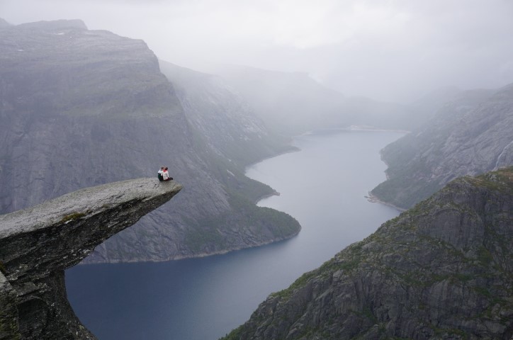 Couple sitting on cliff edge in front of scenery with mountains and rivers.