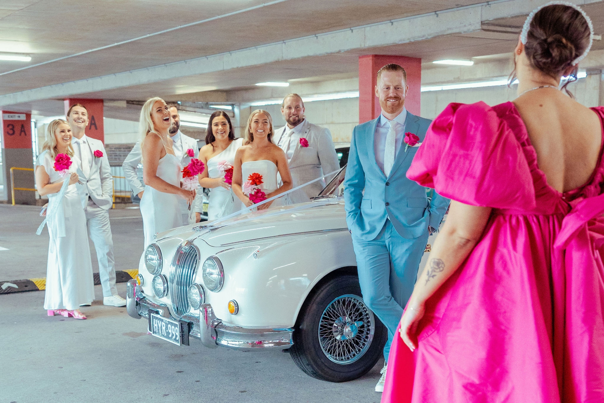Bridesmaids in white hold pink bouquets watching bride walk in with pink wedding dress, groom in blue suit watches his bride