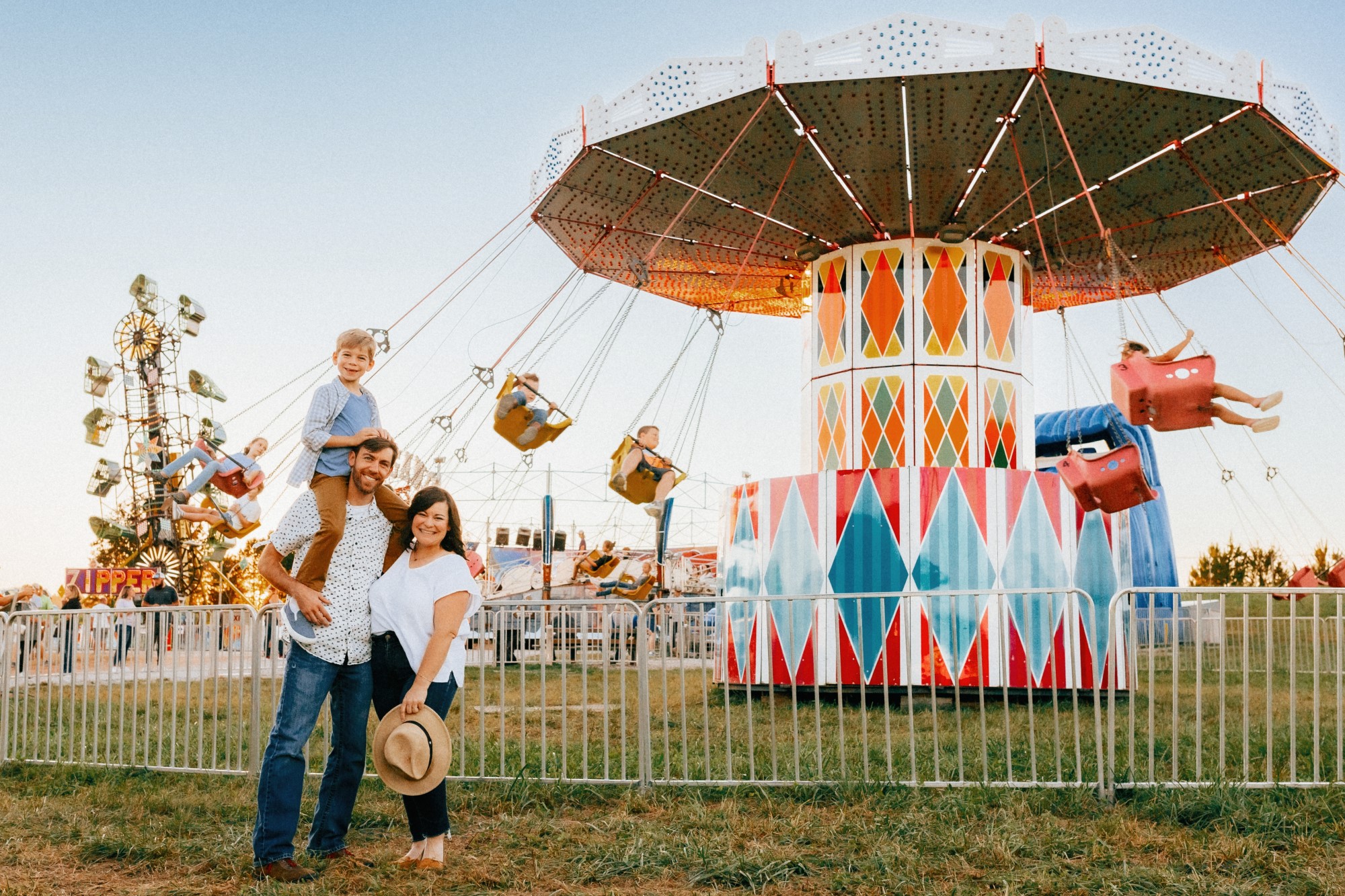 Parents with child on fathers shoulders stand in front of fairground ride at carnival