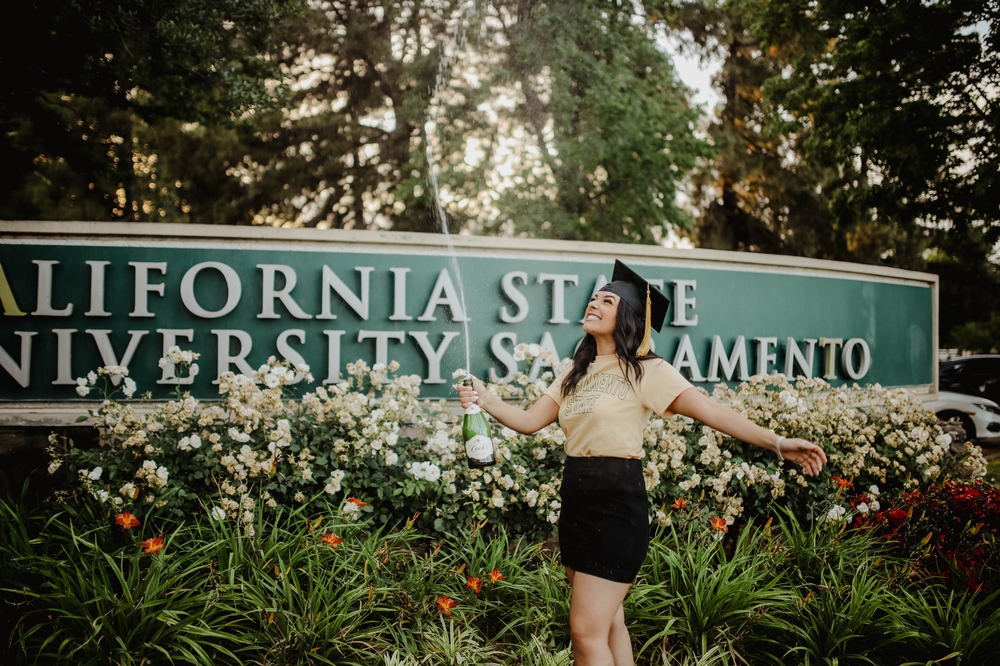 Young woman in casual clothing wearing graduation hat opens champagne in front of university sign
