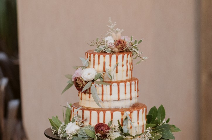 Caramel dripping three tiered wedding cake decorated with white and red flowers in front of brown background