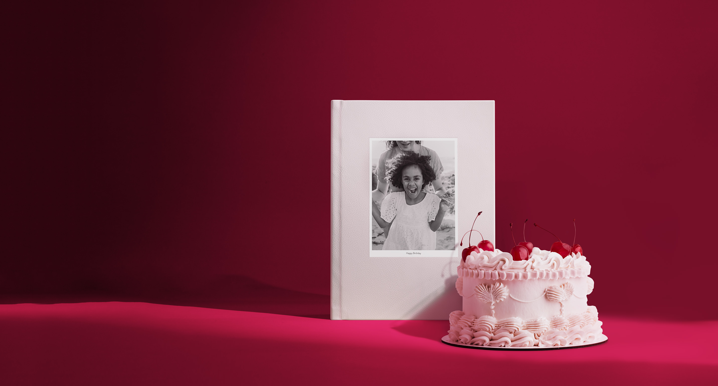 Rosa Porträt-Premium-Fotobuch neben einer rosa Torte mit Kirschen auf rotem Hintergrund