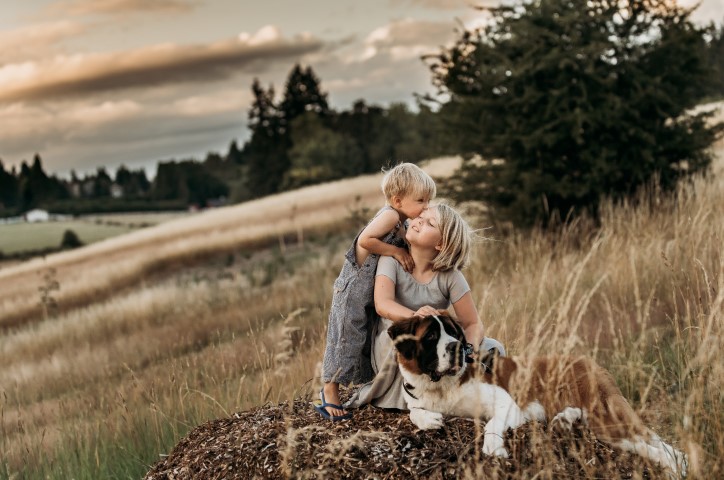 Young girl and her brother sit on bark in dried grass field with their large mountain dog