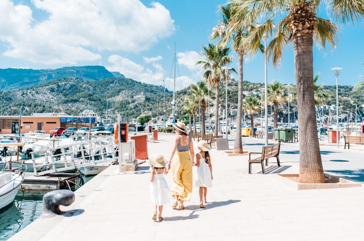 Mother with two children walk on sunny beach with boats lined on wharf