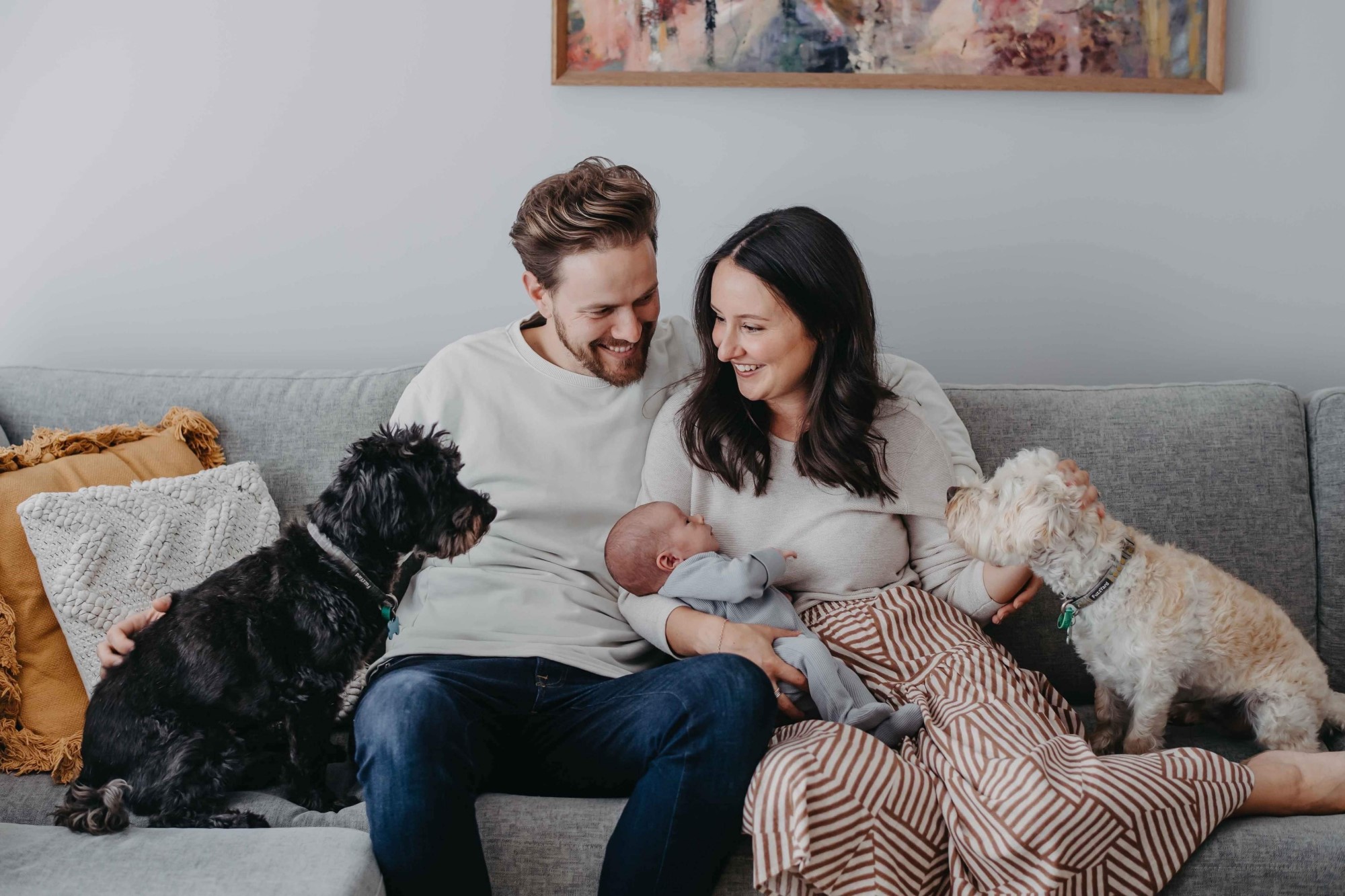 Parents sit on couch smiling holding baby with dogs on either side