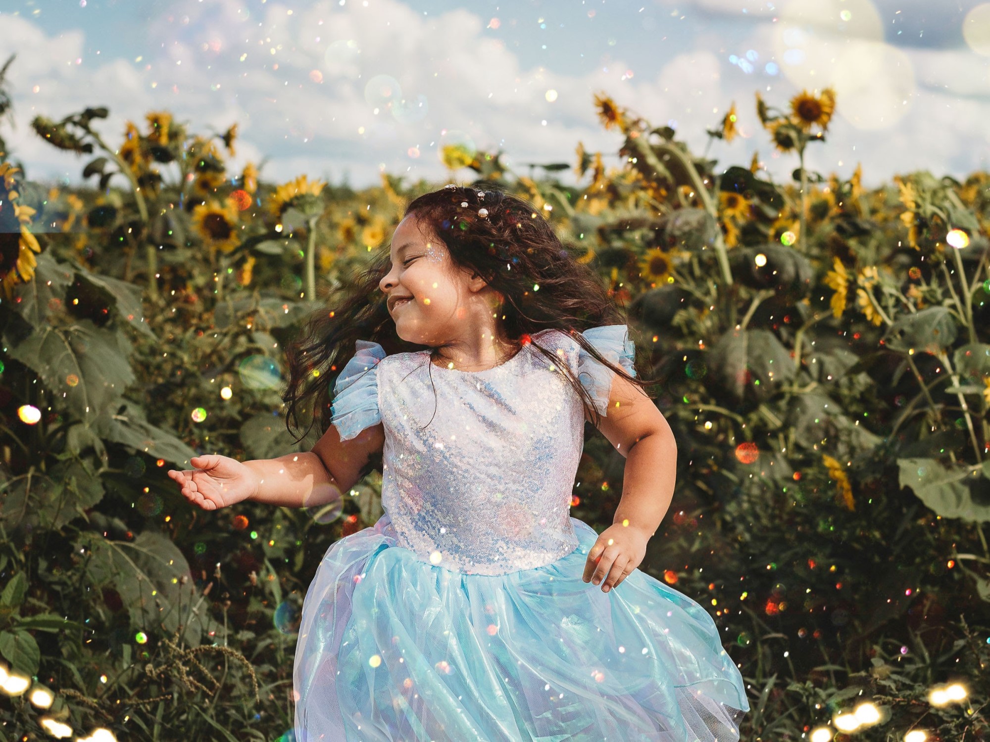 Young girl in blue fairy dress dancing in sunflower field under blue sky with glitter everywhere