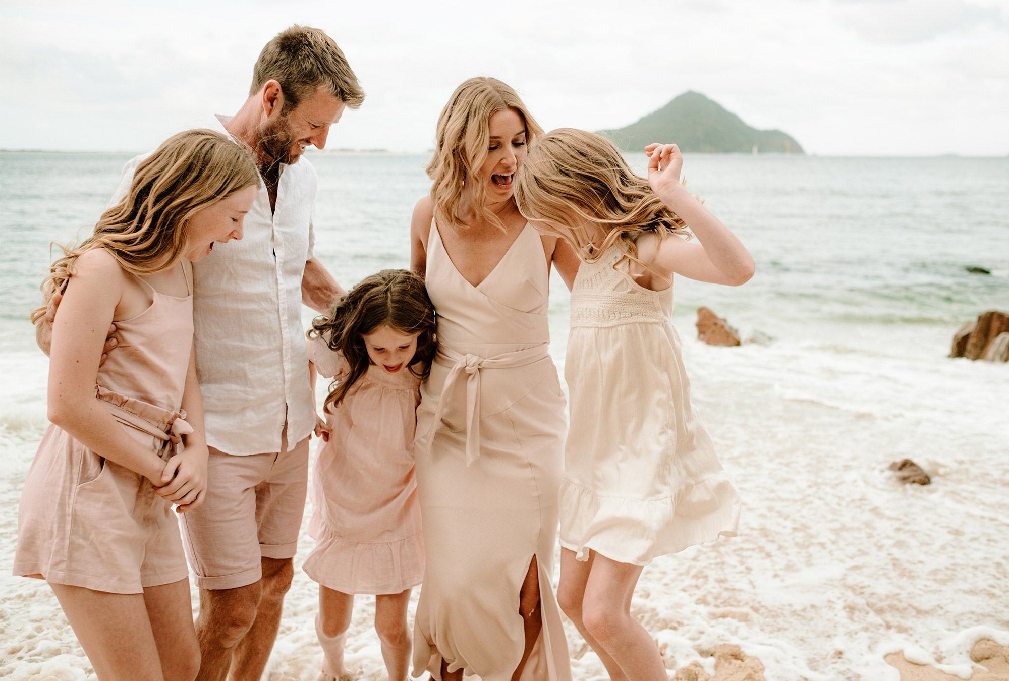 Family of five in matching light pink outfits are startled at crashing wave they are standing in on beach shore.