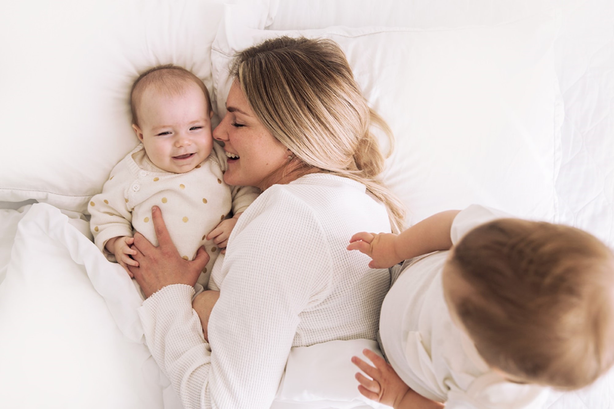 Mother lying in white bed with two babies smiling together