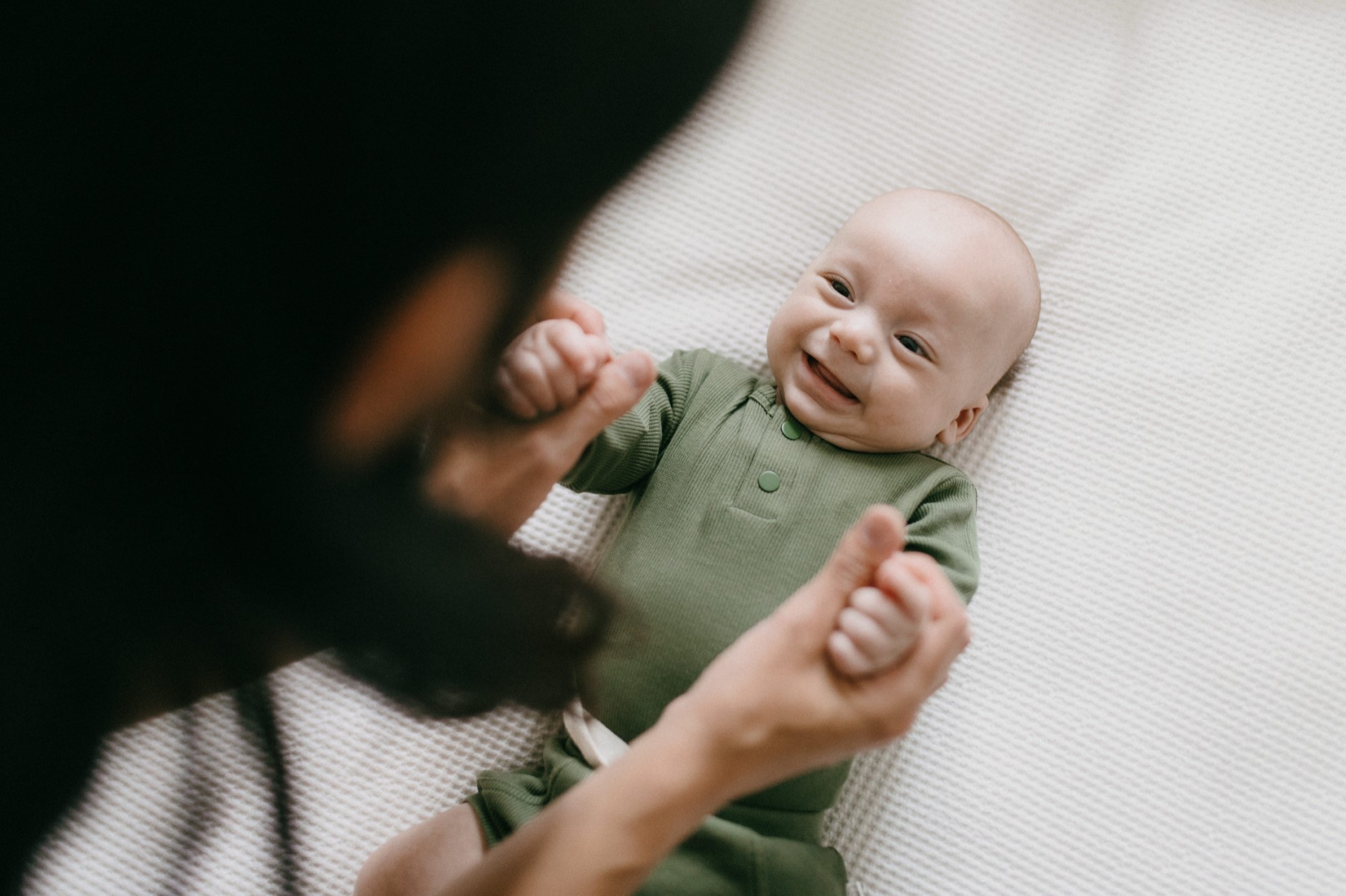 Baby lying on white sheets wearing green onesie and smiling with dark haired mother in foreground