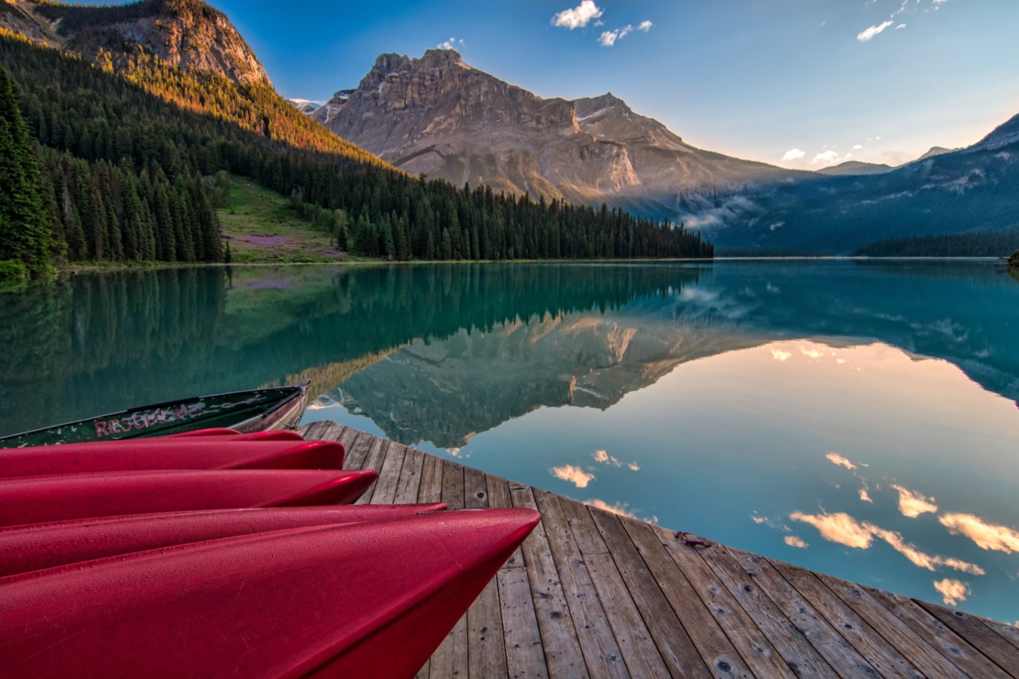 Kayaks on dock next to reflective lake surrounded by mountains and forest under blue sky near sunset