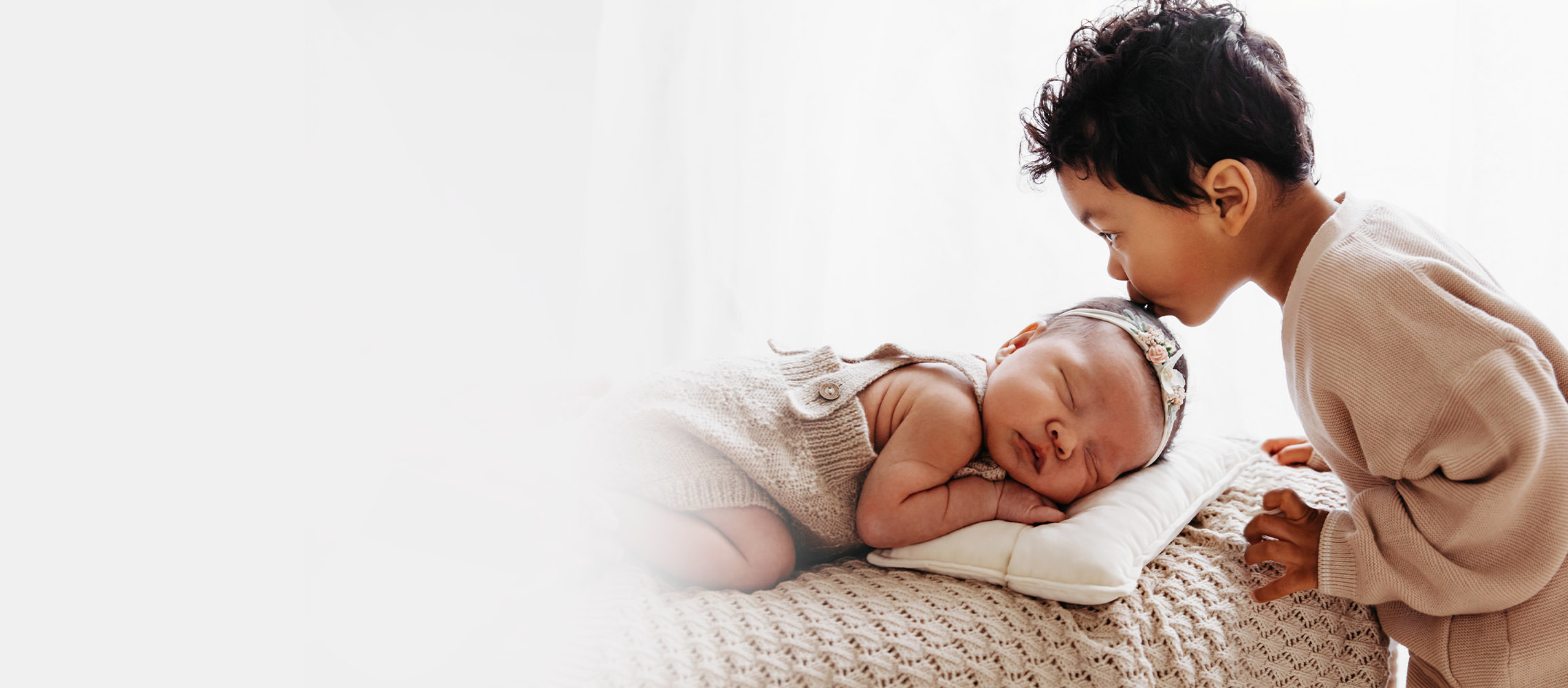 Toddler kisses newborn baby's head as they sleep on pillow