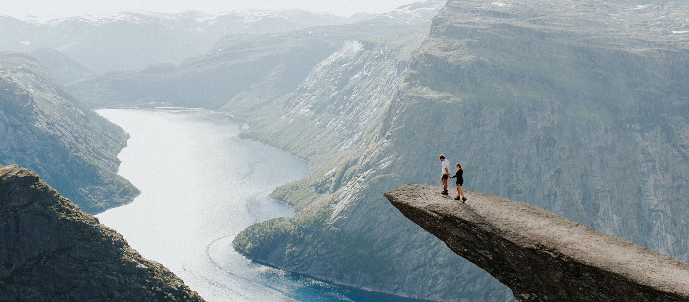 Couple hold hands to the edge of a cliff overlooking river and mountain ravine