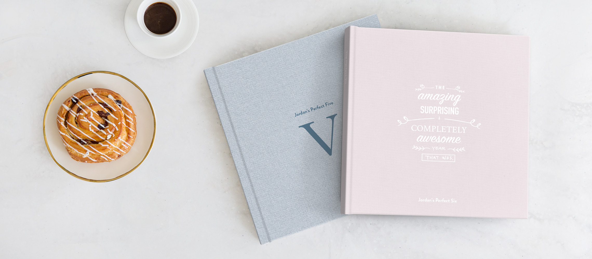 Dois álbuns de fotos de aniversário em uma mesa ao lado de um café e um croissant.