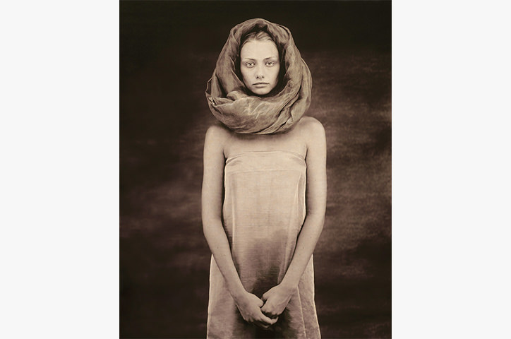 Pale woman in beige wrapped outfit clasping hands standing against dark background