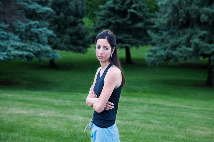 Dark haired woman crosses arms standing in lush green park field with surrounding trees