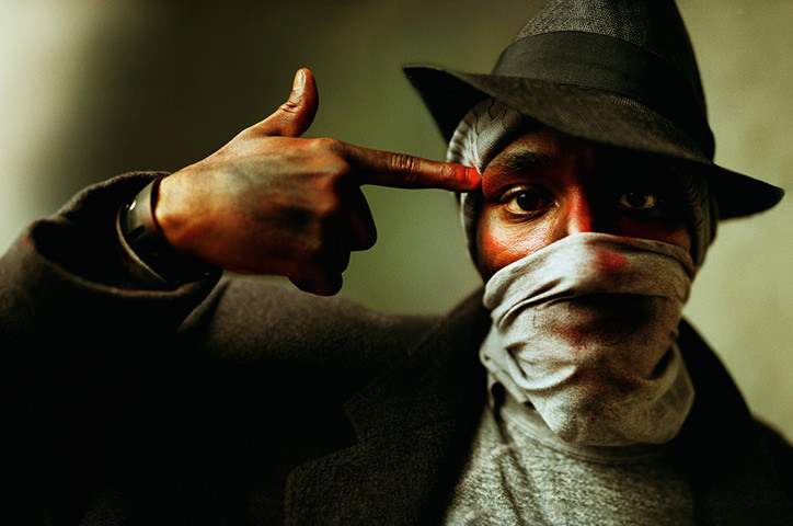 Man covering mouth with wrap wearing dark outfit points to his head