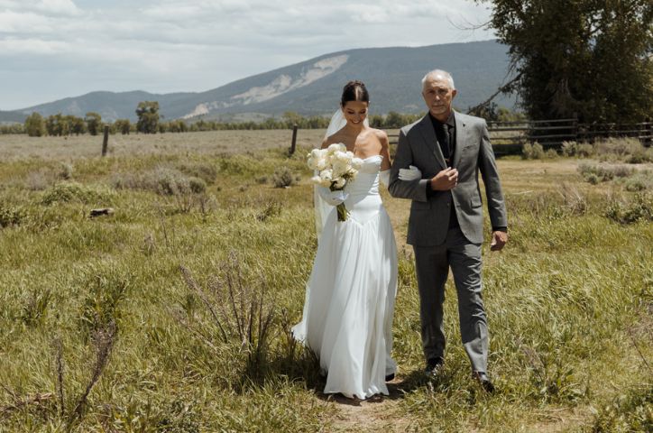 Smiling bride walking through farm during wedding ceremony with her father on her arm.
