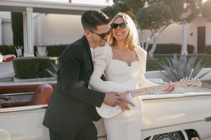 Engaged couple smile and play a white guitar together leaning on vintage car in California