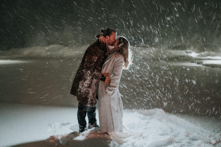 Newlywed couple kiss under winter snowfall at night with spotlight