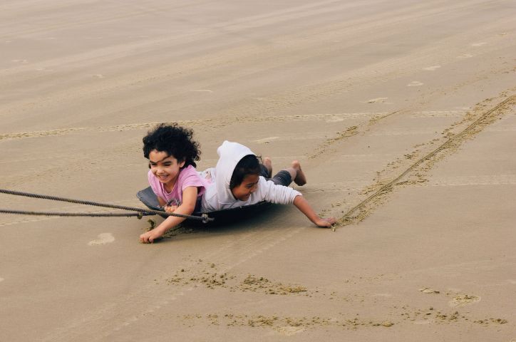 Two children smiling lying on a board sliding across beach