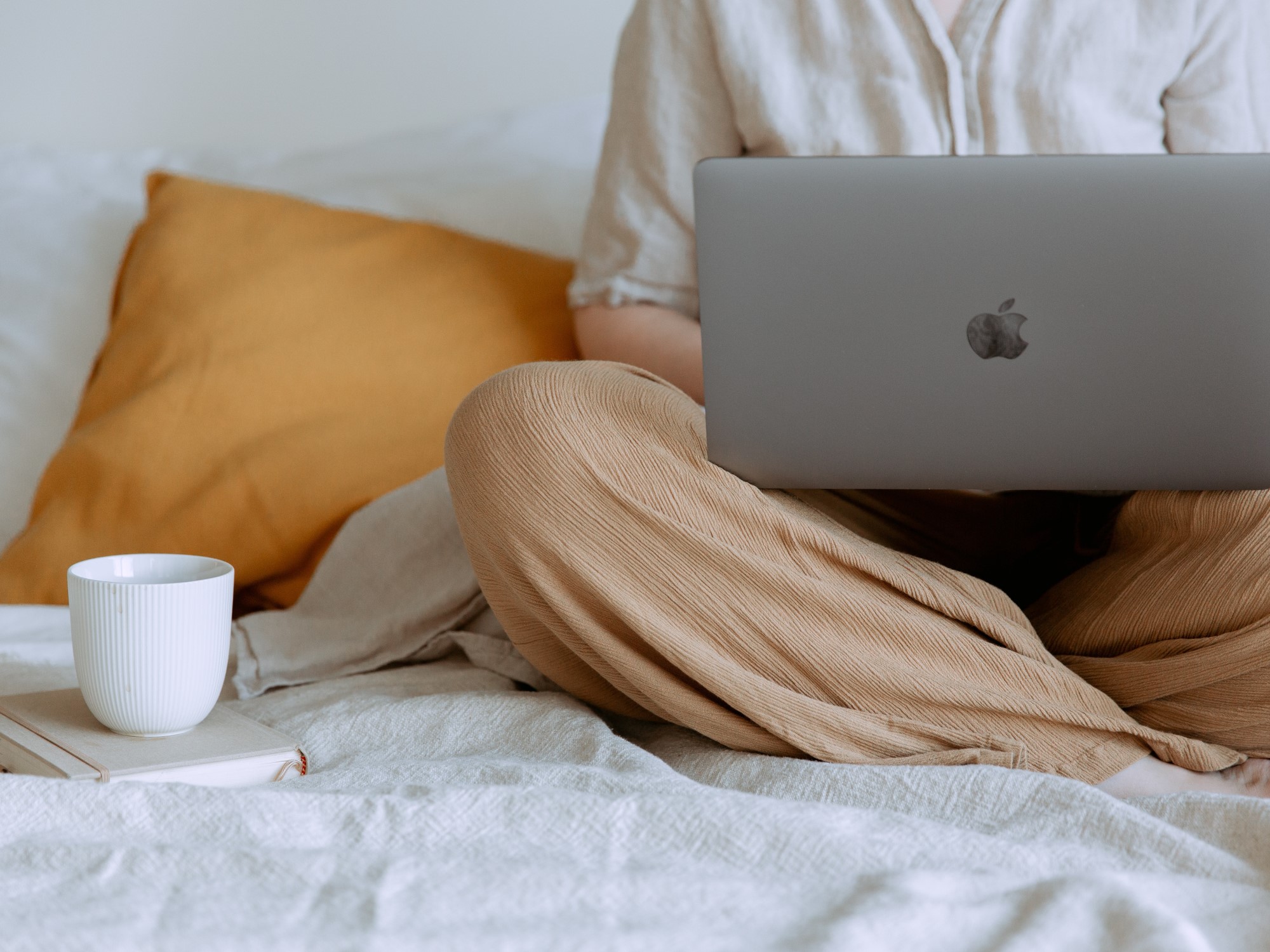 Person holding laptop on their knees while sitting on bed with yellow pillow white sheets and white coffee mug on book