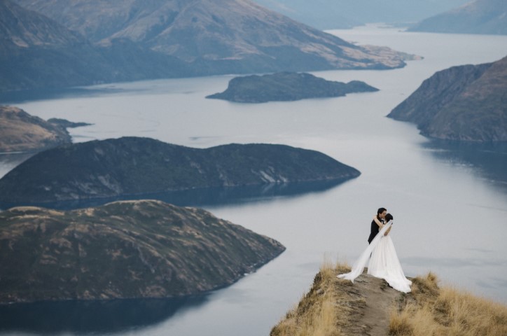 Newlywed couple stand on mountaintop edge overlooking lake while kissing.