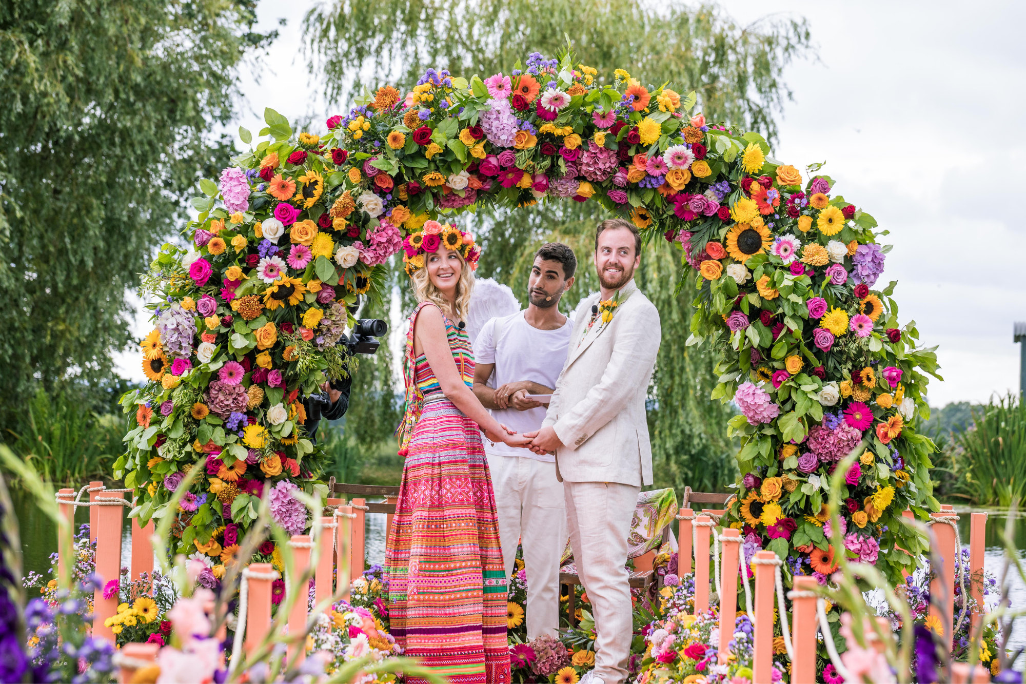 Bride wears bold pink wedding outfit and holds hands with groom at end of colorful and flowery aisle