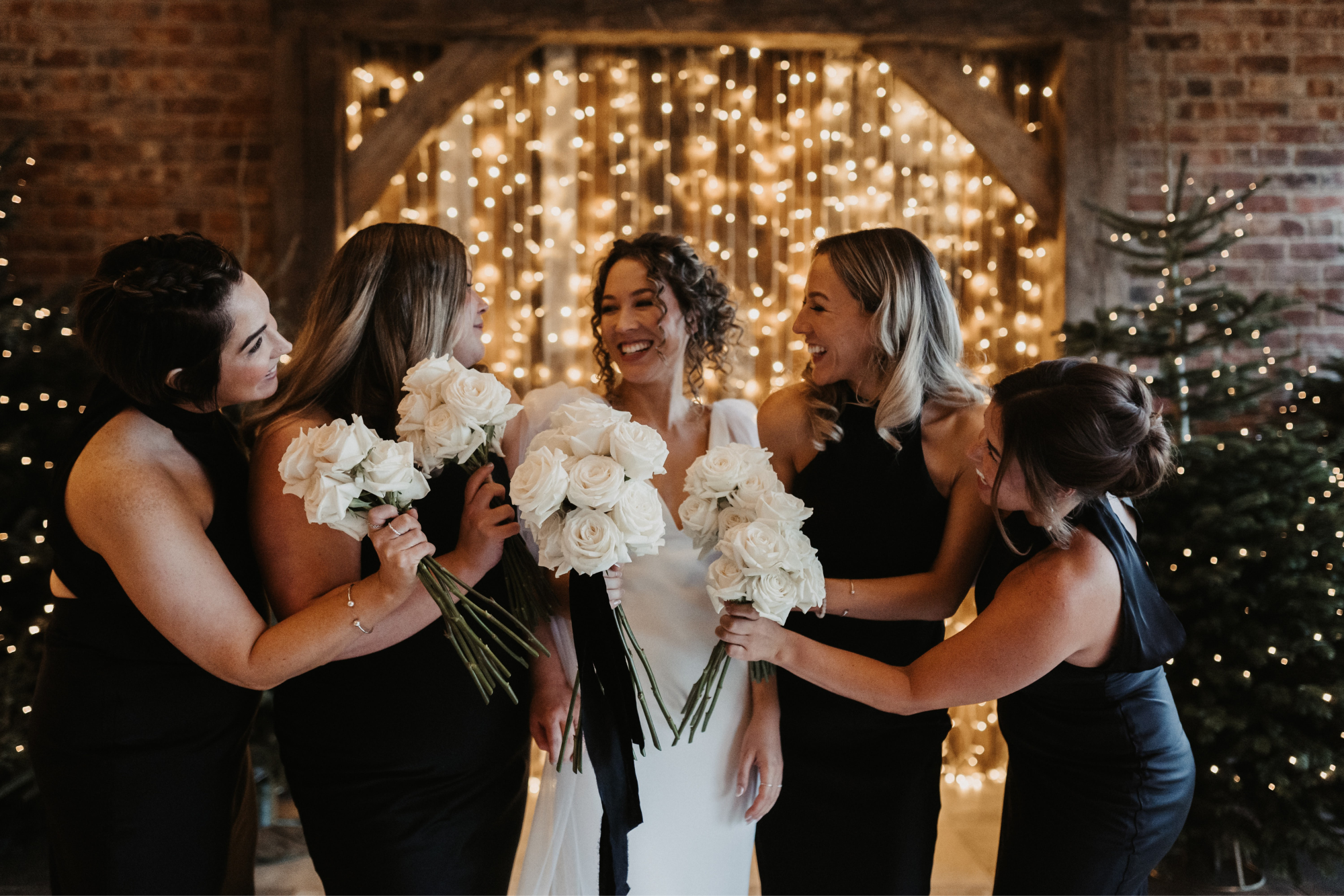 Bridal party wearing black dresses stand in front of fairy lights on wall with bride