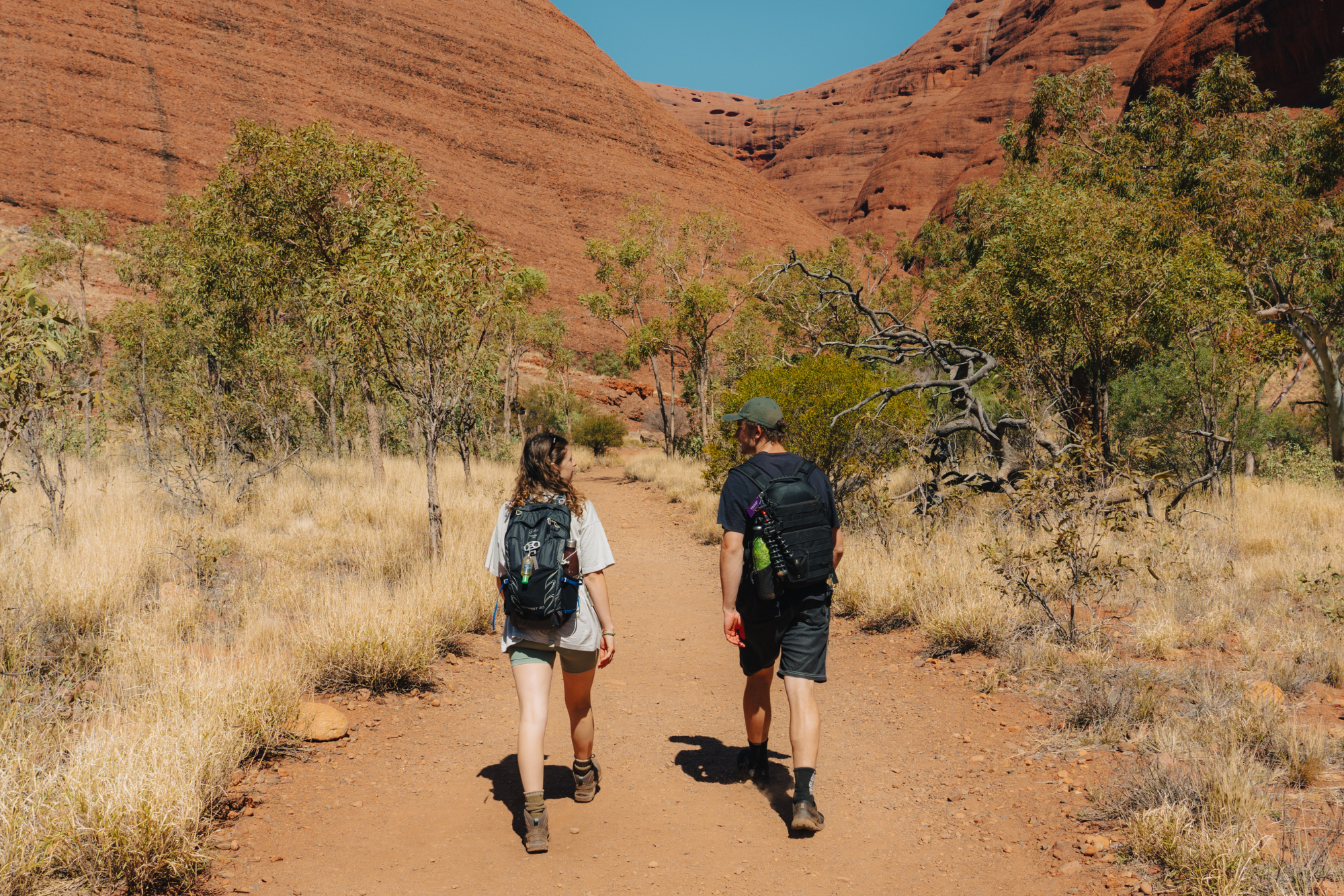 Two hikers walk down dirt path towards red Ayers rock in Australia