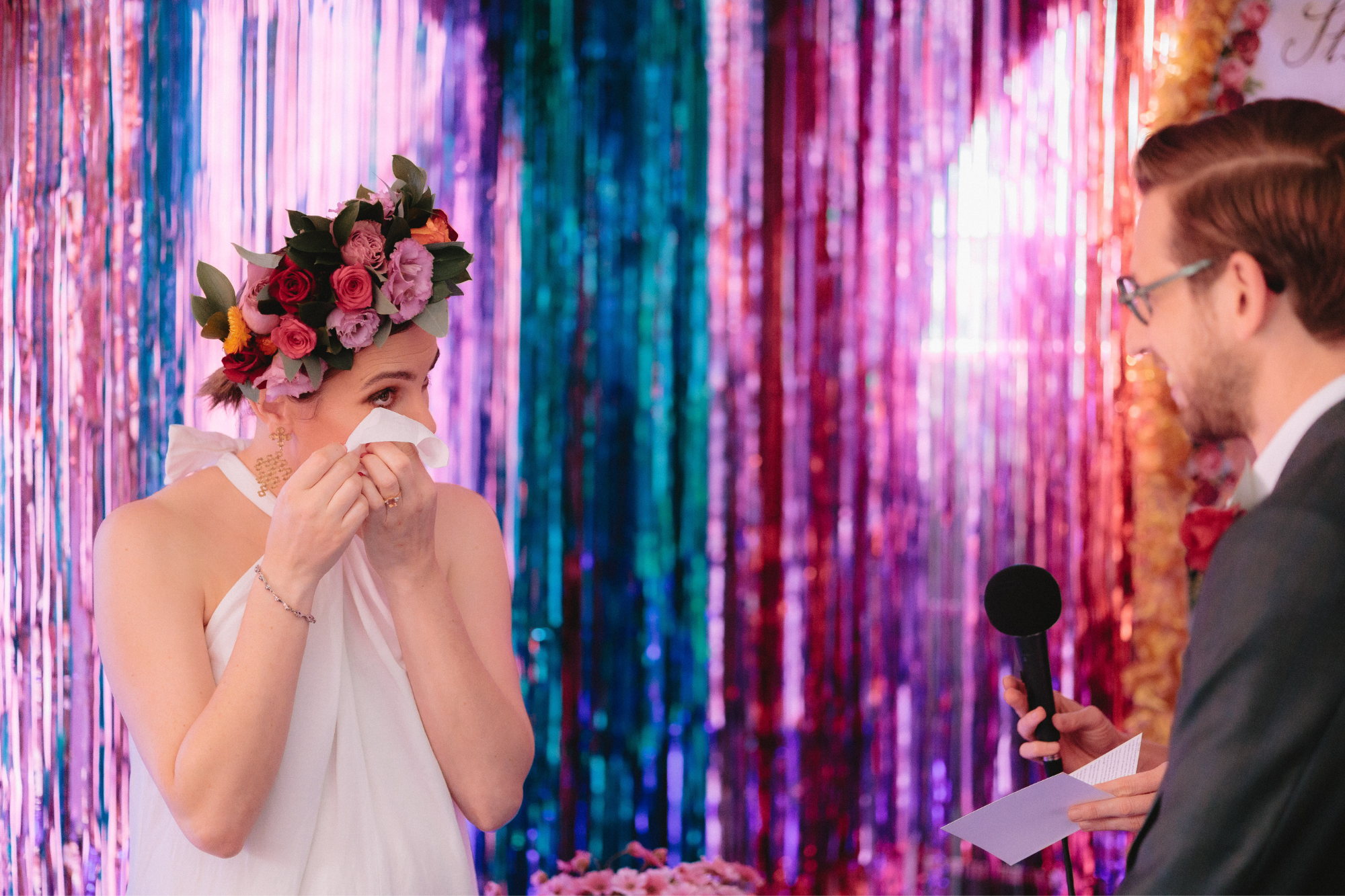 Bride uses tissue to wipe tears and wears flower crown at end of aisle with metallic streamers on wall