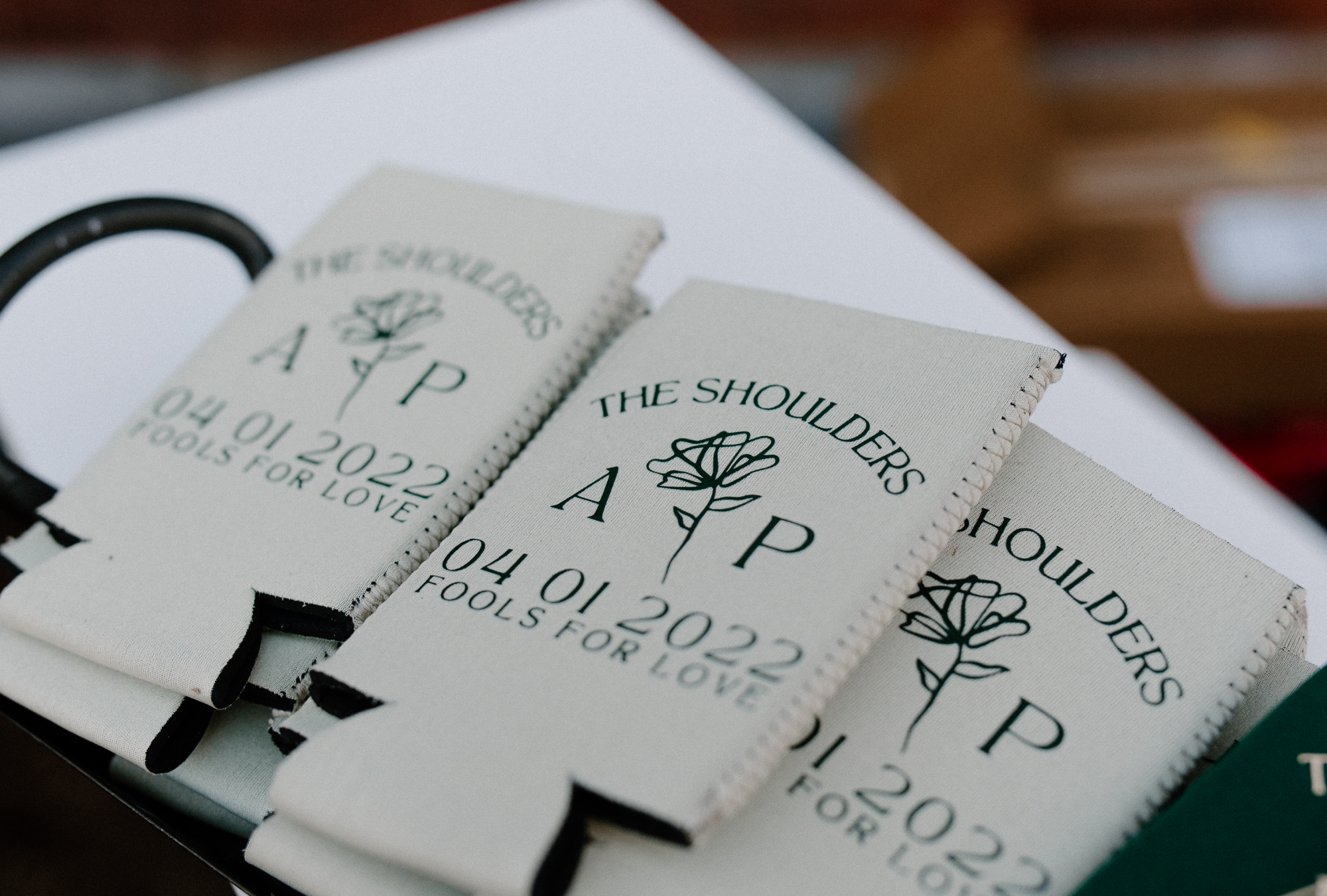 Custom white koozies labelled "The Shoulders" with initials on them, laid on white table