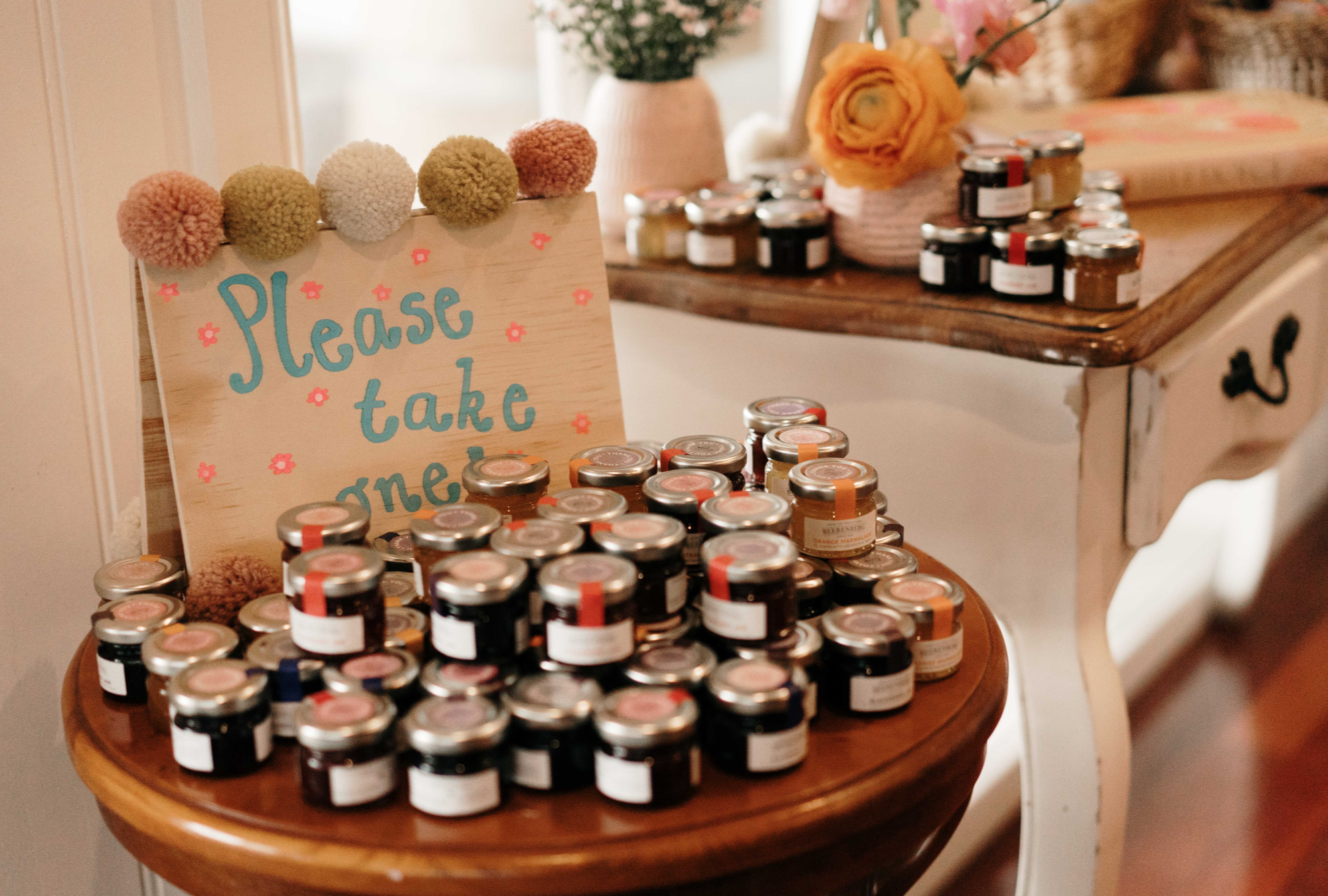 "Please take one!" blue writing on a wooden sign behind stacks of personalized labelled jams and jellies and other preserves