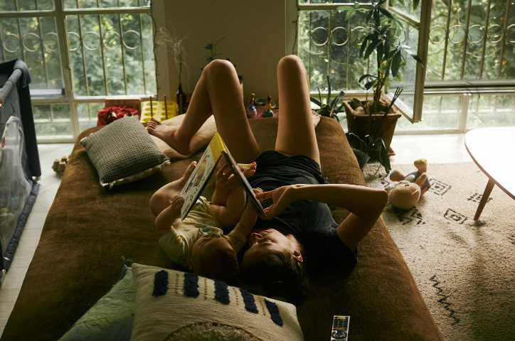 Father lies on back with toddler son on couch reading picture book