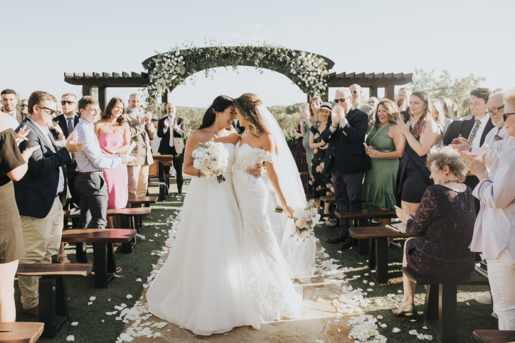 Two brides walk down the wedding aisle together wearing white dresses